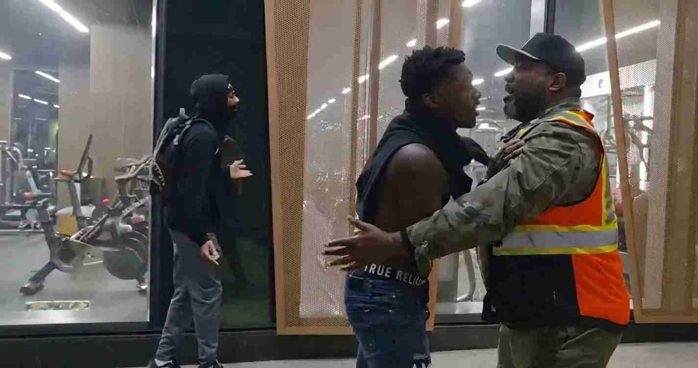 Security guard in an orange-vested uniform holds back a shirtless man as a bystander in a black hoodie approaches from the left near gym glass walls and equipment.