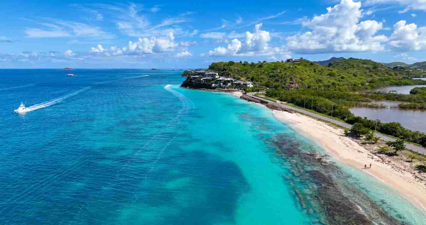 Aerial view of a tropical beach with turquoise water, white sand, and a green hillside dotted with houses along the shore.