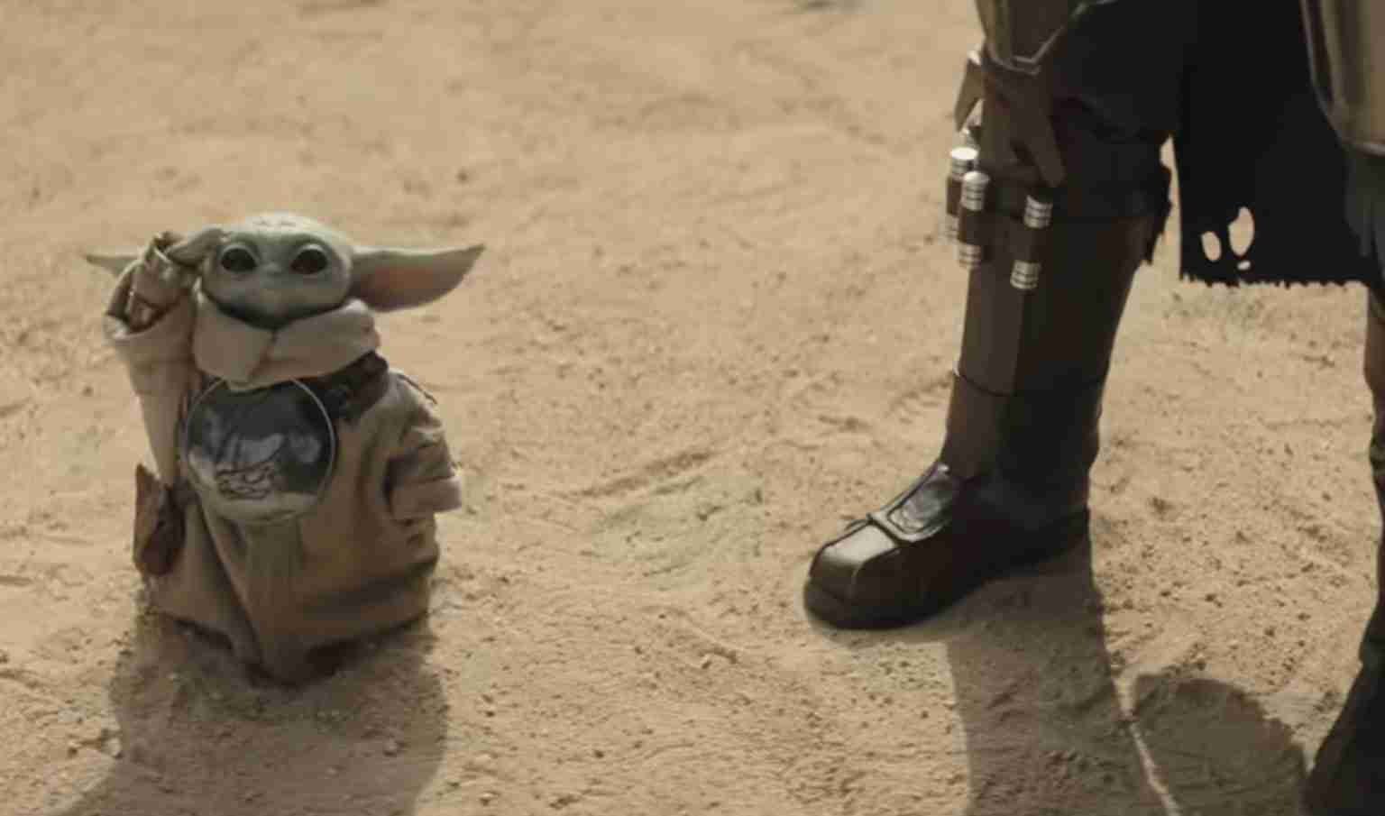 Baby Yoda (Grogu) figurine stands in a sandy desert next to a Mandalorian’s armored boot.