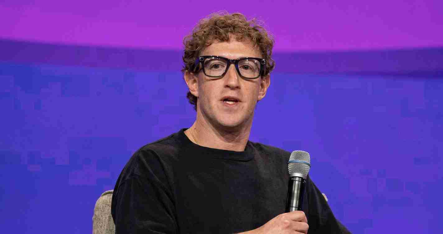 Man with curly hair and glasses holding a microphone on a purple-stage backdrop, mid-speech.