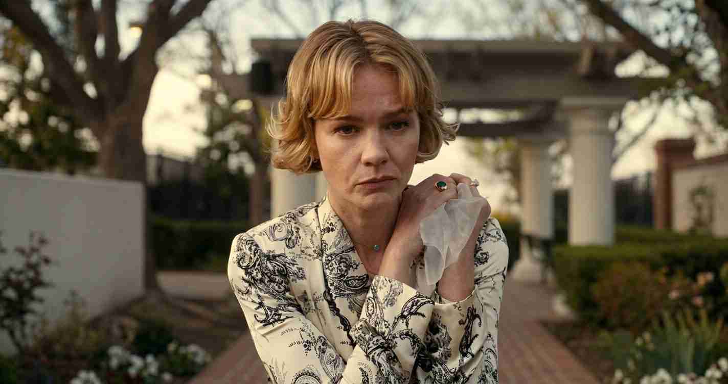 Woman with short blonde hair in a paisley-patterned blouse, clasping a scarf and looking somber outdoors in a garden path setting.