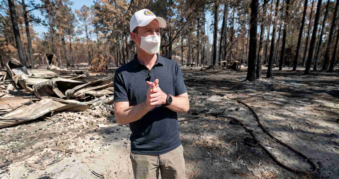 Man wearing a white mask and cap stands in a scorched forest with debris and charred ground behind him, gesturing with his hands.