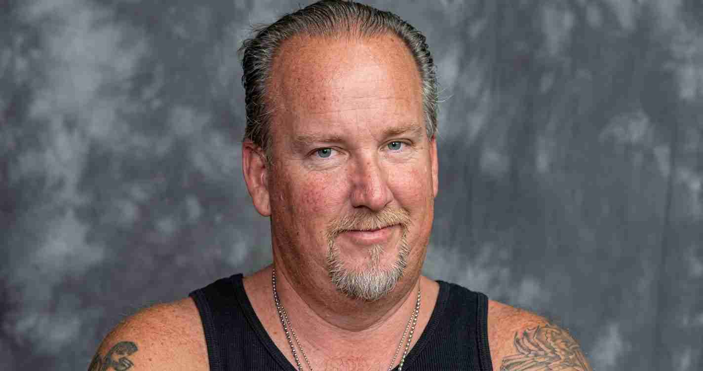 Portrait of a middle-aged man with a goatee wearing a black sleeveless shirt, with tattoos on his arm, against a gray studio backdrop.