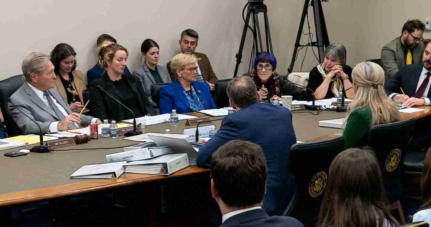Group of professionals seated around a conference table in a formal meeting with microphones, folders, and water bottles on the table.