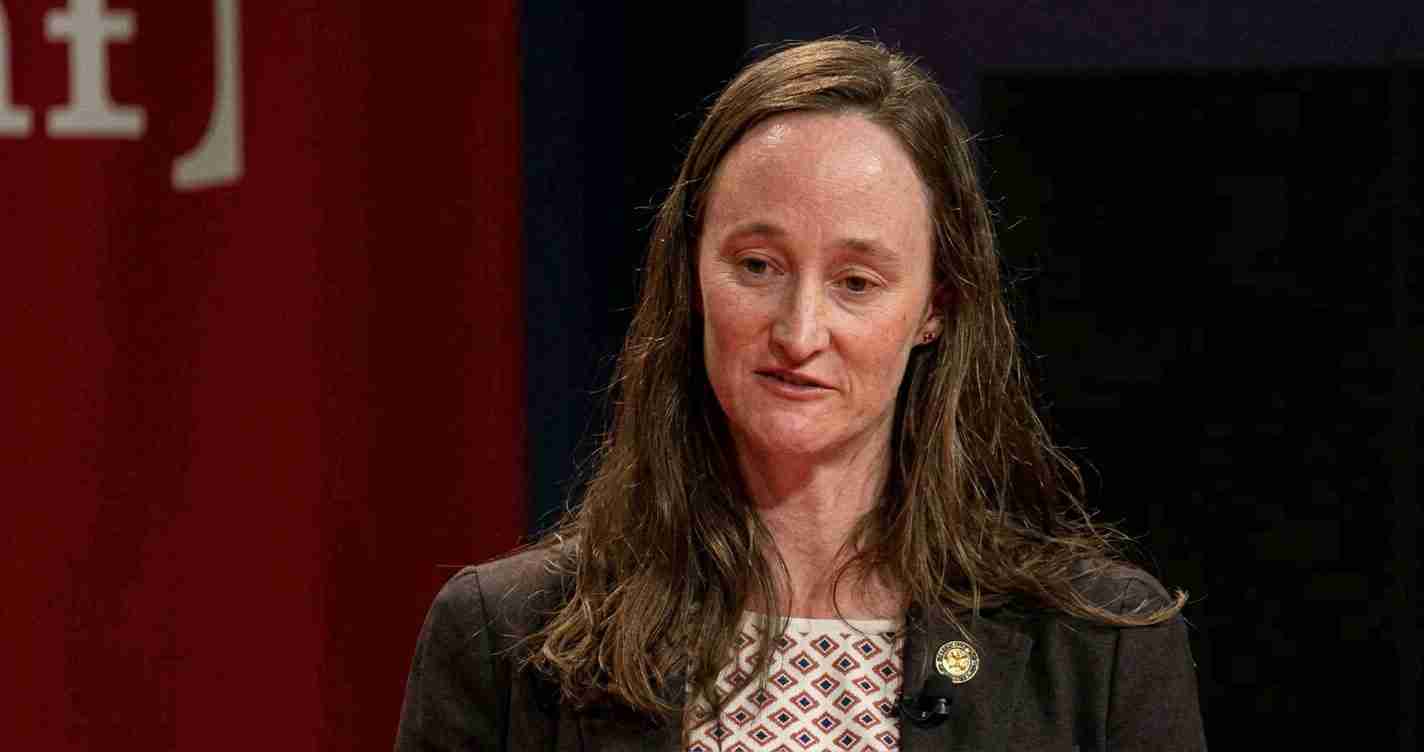Woman with long brown hair and blazer speaking on stage, wearing a patterned blouse and a small lapel pin on her left lapel.