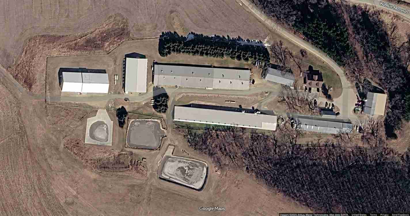 Aerial view of an industrial complex with long rectangular buildings, circular and rectangular basins, and surrounding fields near a road.