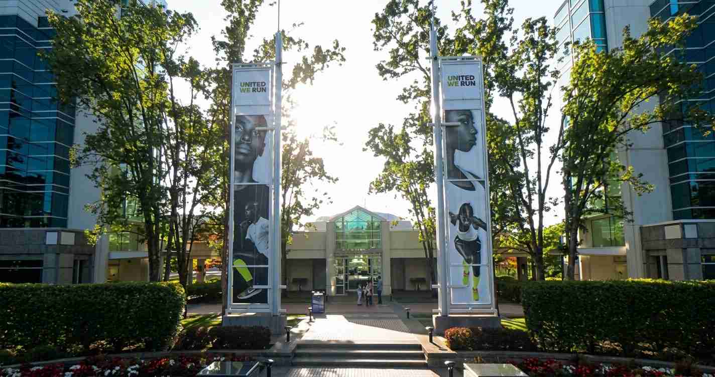 Entrance to a modern campus with two vertical banners reading United We Run, flanked by trees and shrubs.