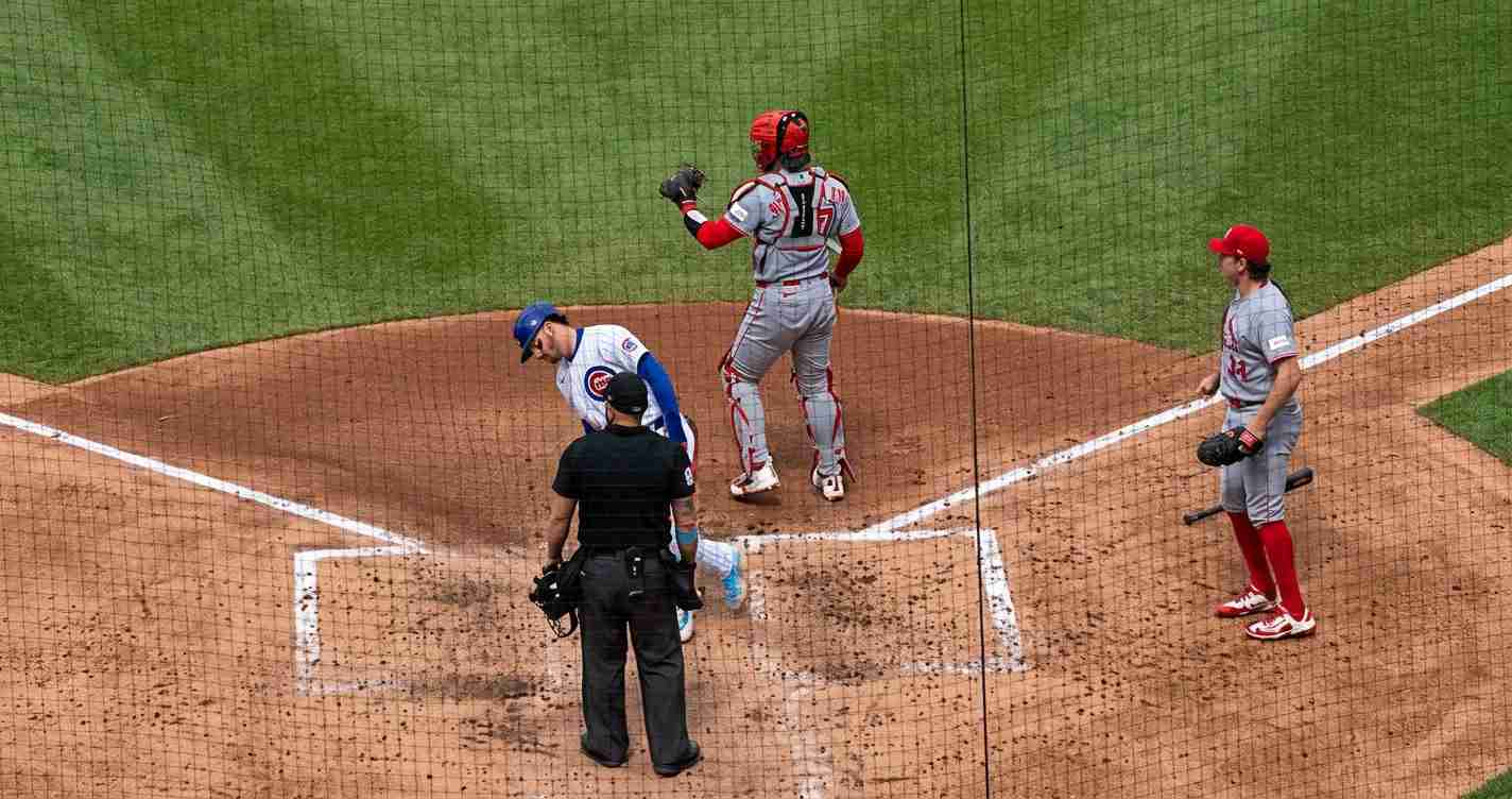 Baseball game at home plate with an umpire, a Cubs batter in white/blue, and a gray-uniform catcher with a red helmet near the plate.