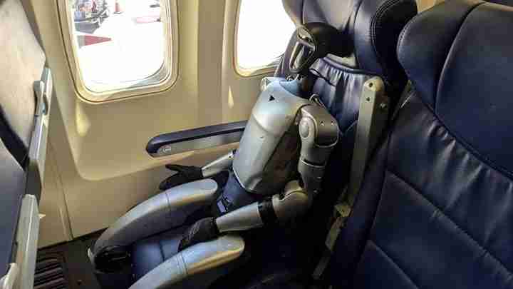 Humanoid robot seated in an airplane seat by a window with blue leather cushions and folded arms.