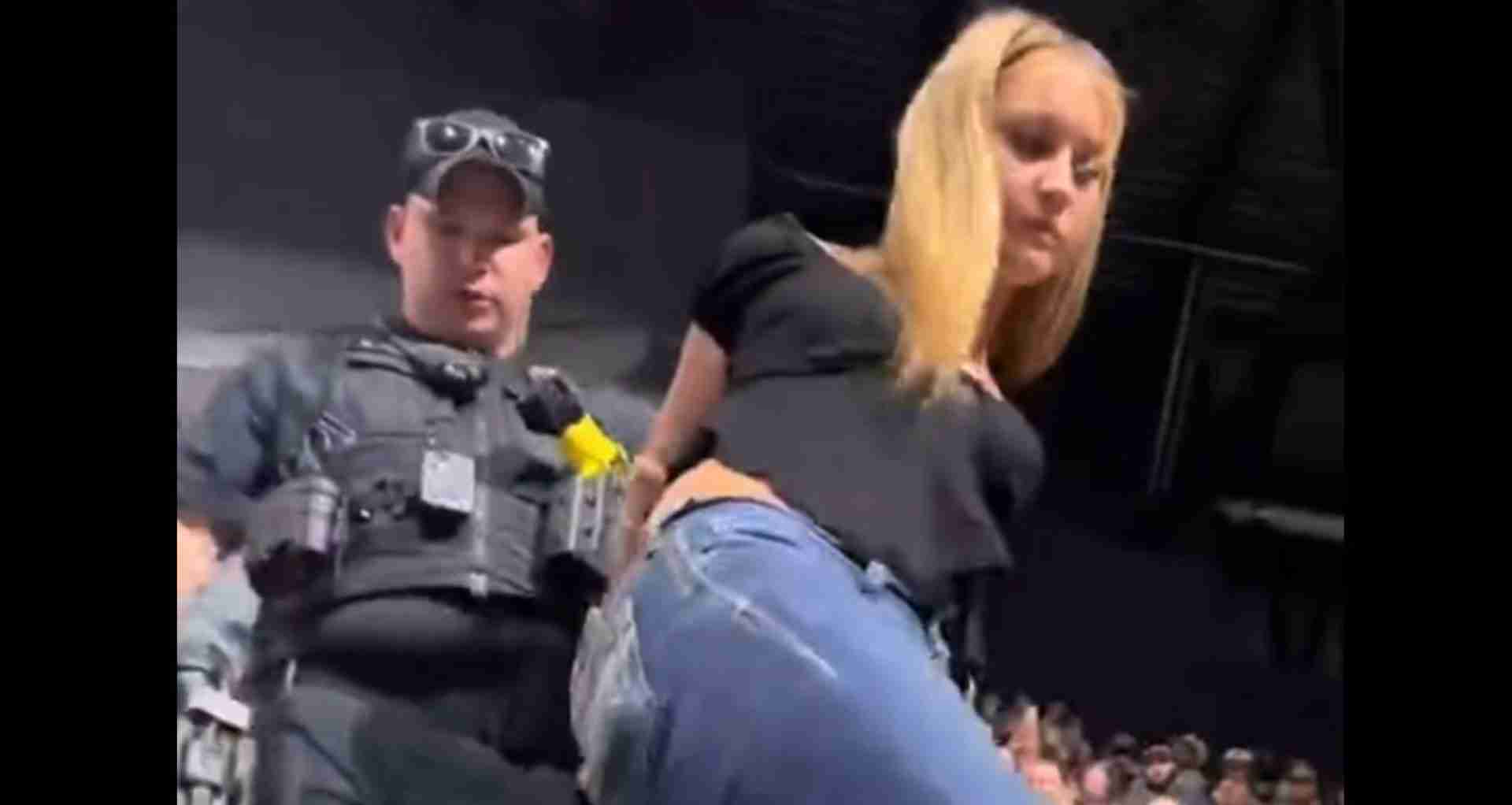 Security guard in a tactical vest stands beside a blonde performer bowing toward a cheering crowd on a dimly lit stage.