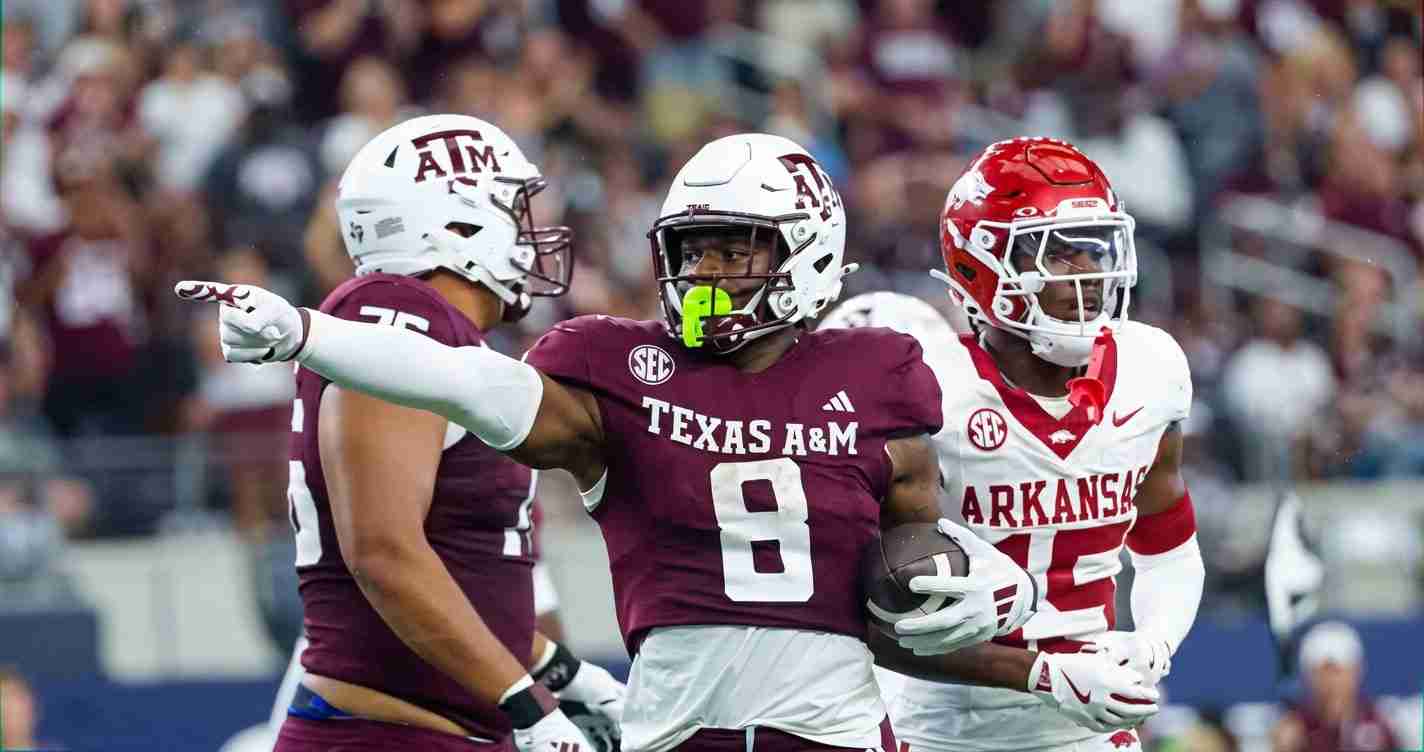 Texas A&M running back in maroon jersey (#8) carries the football while an Arkansas defender in white pursues on the field.
