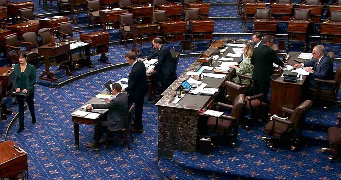 Legislative chamber with desks and laptops; lawmakers stand and sit around a marble podium on a blue-patterned floor.