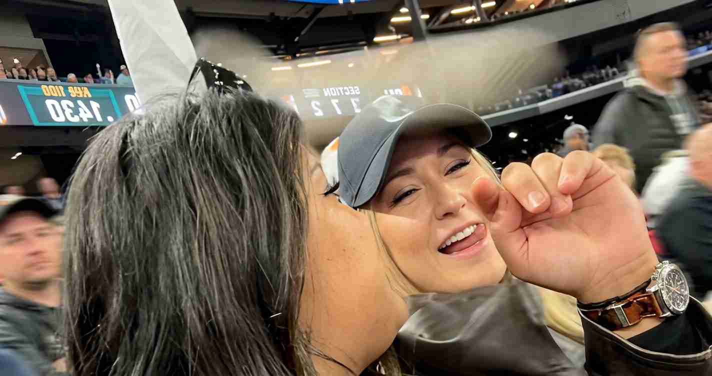 Two friends at a sports stadium share a kiss on the cheek; one wears a gray cap and a wristwatch.