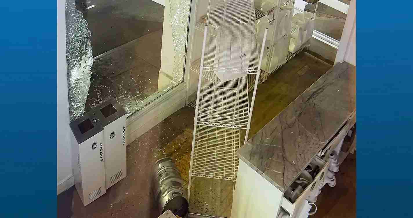 Interior of a store after a break-in: shattered window, toppled metal display rack, and scattered debris on the floor near white electrical cabinets labeled GE/SYNMOC? (damage evident).
