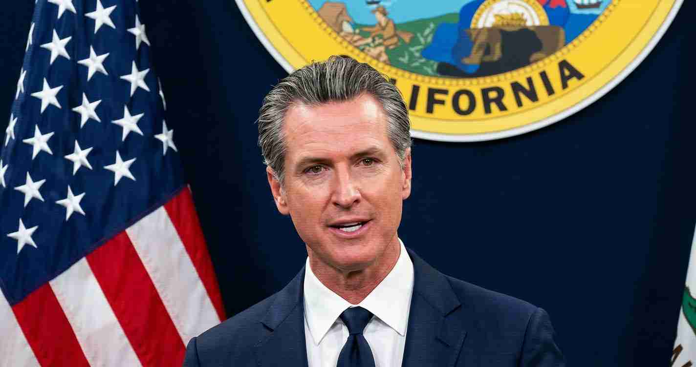 Male politician in a navy suit speaking at a press conference, with the U.S. flag and California state seal visible behind him.