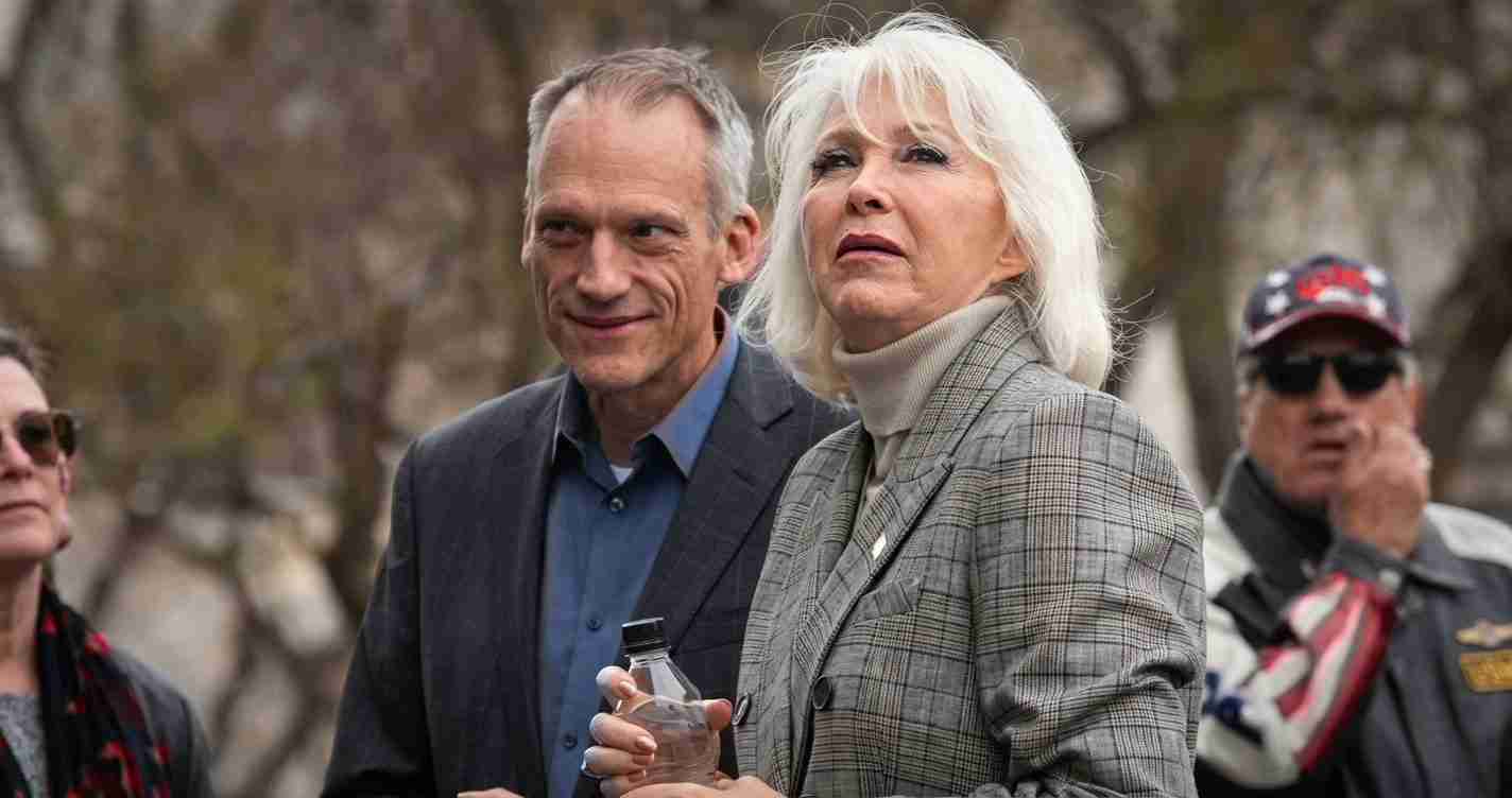 Two older adults outdoors; woman in a gray plaid blazer holds a water bottle while a man in a blue shirt stands beside her.