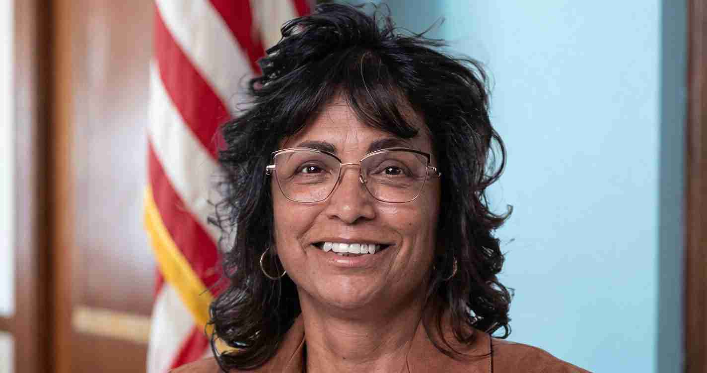 Smiling woman with dark, wavy hair and glasses, standing in front of an American flag.