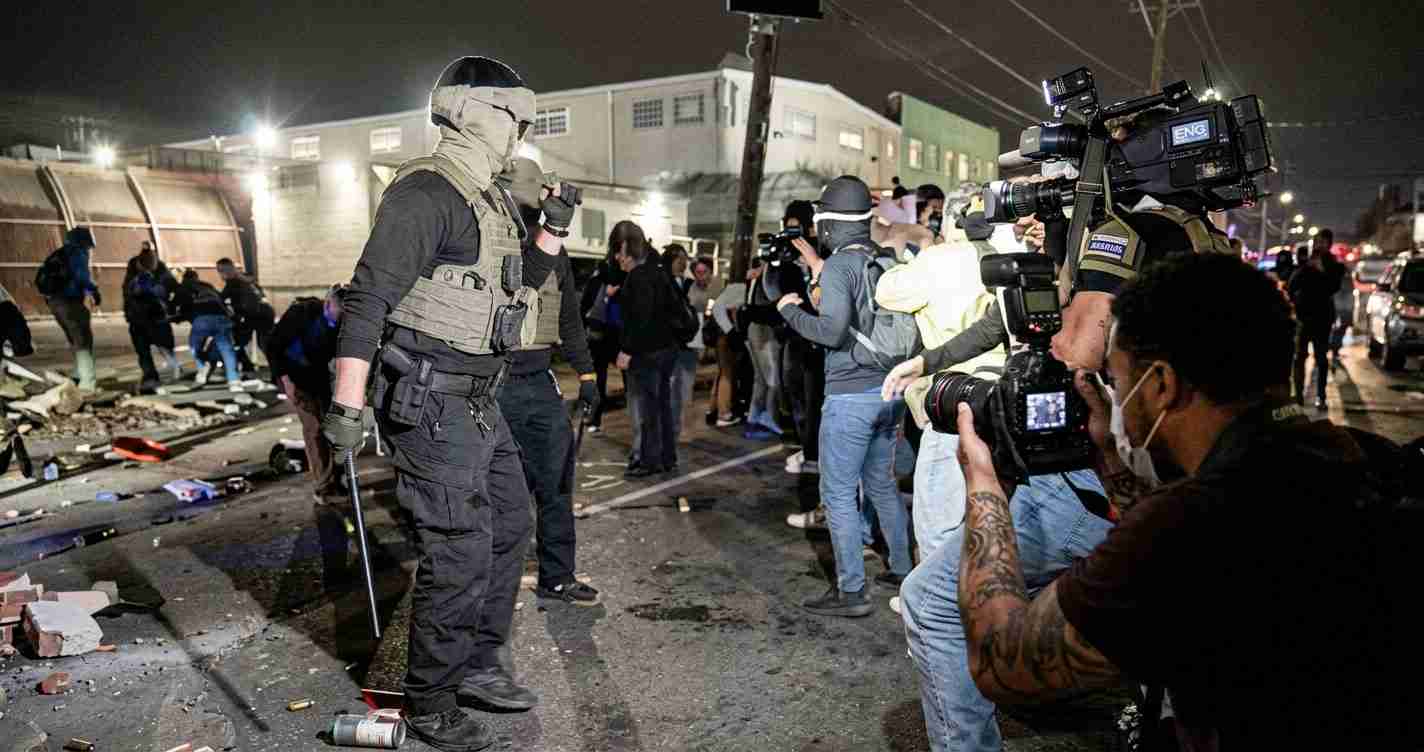 Armed officer in tactical gear stands between a crowd and media on a nighttime street scene, with debris on the ground and photographers nearby