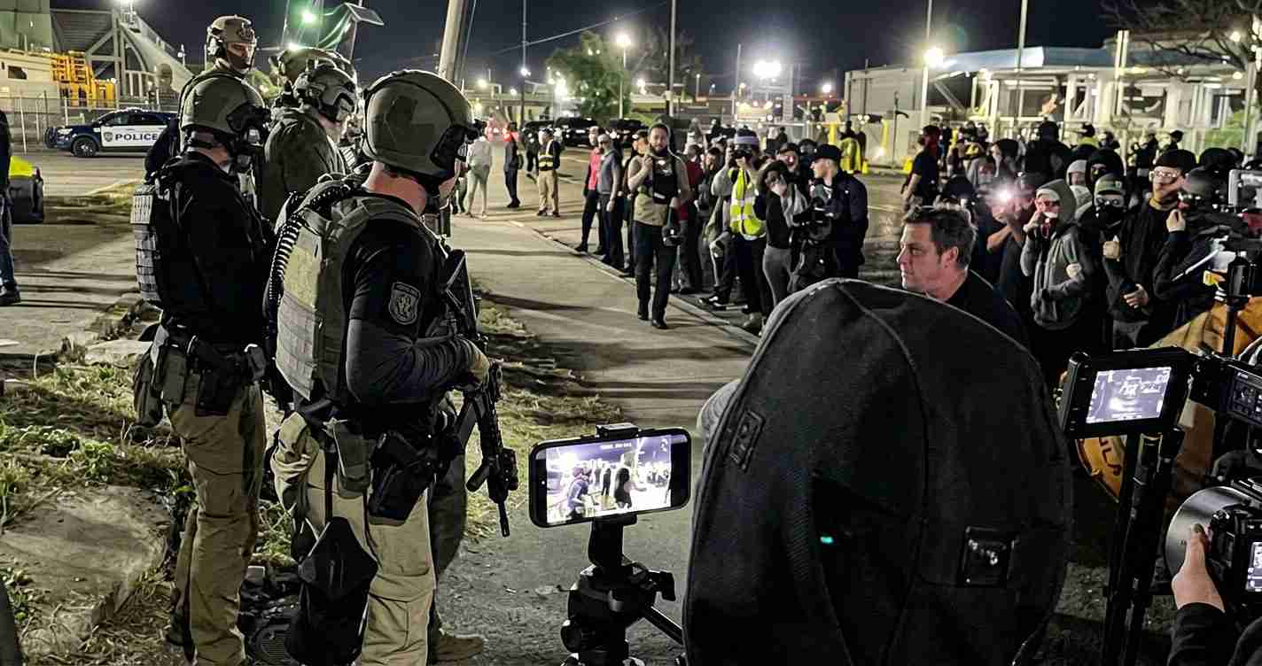 Tactical police officers in armor stand before a crowd at night as protesters and media document the scene.