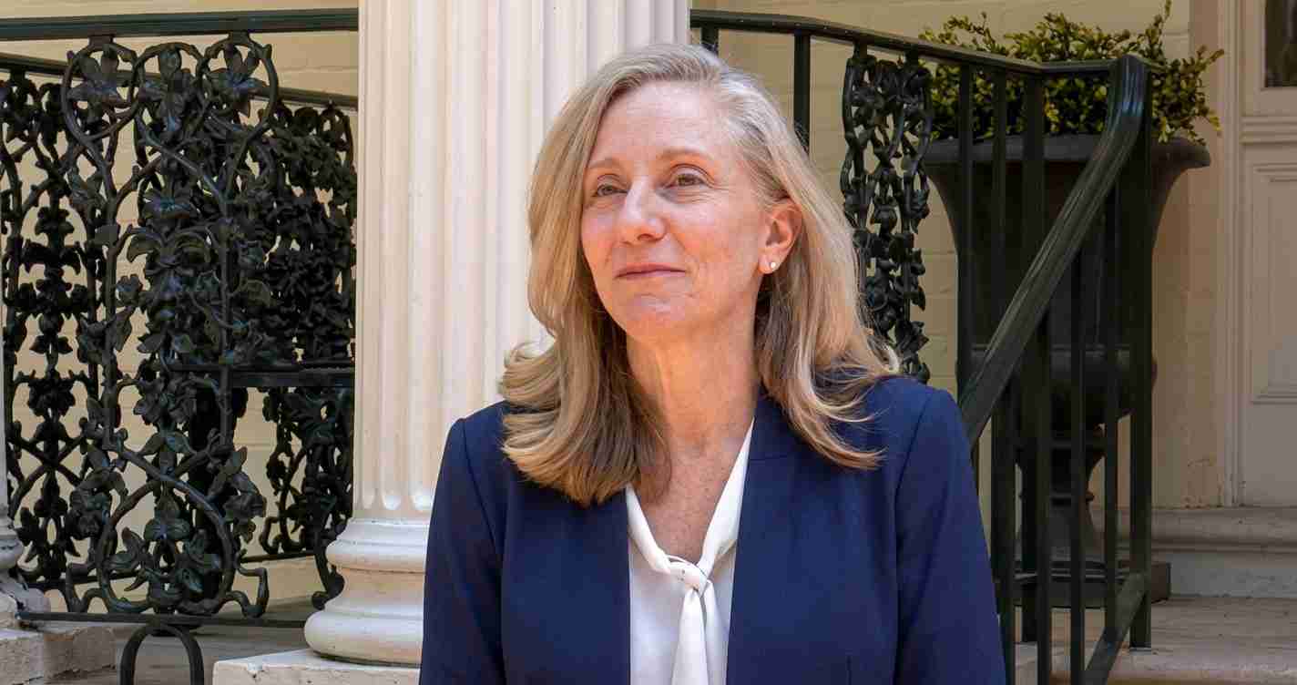 Portrait of a woman with shoulder-length blonde hair in a navy blazer and white blouse, standing on a stone porch with decorative iron railing behind her.