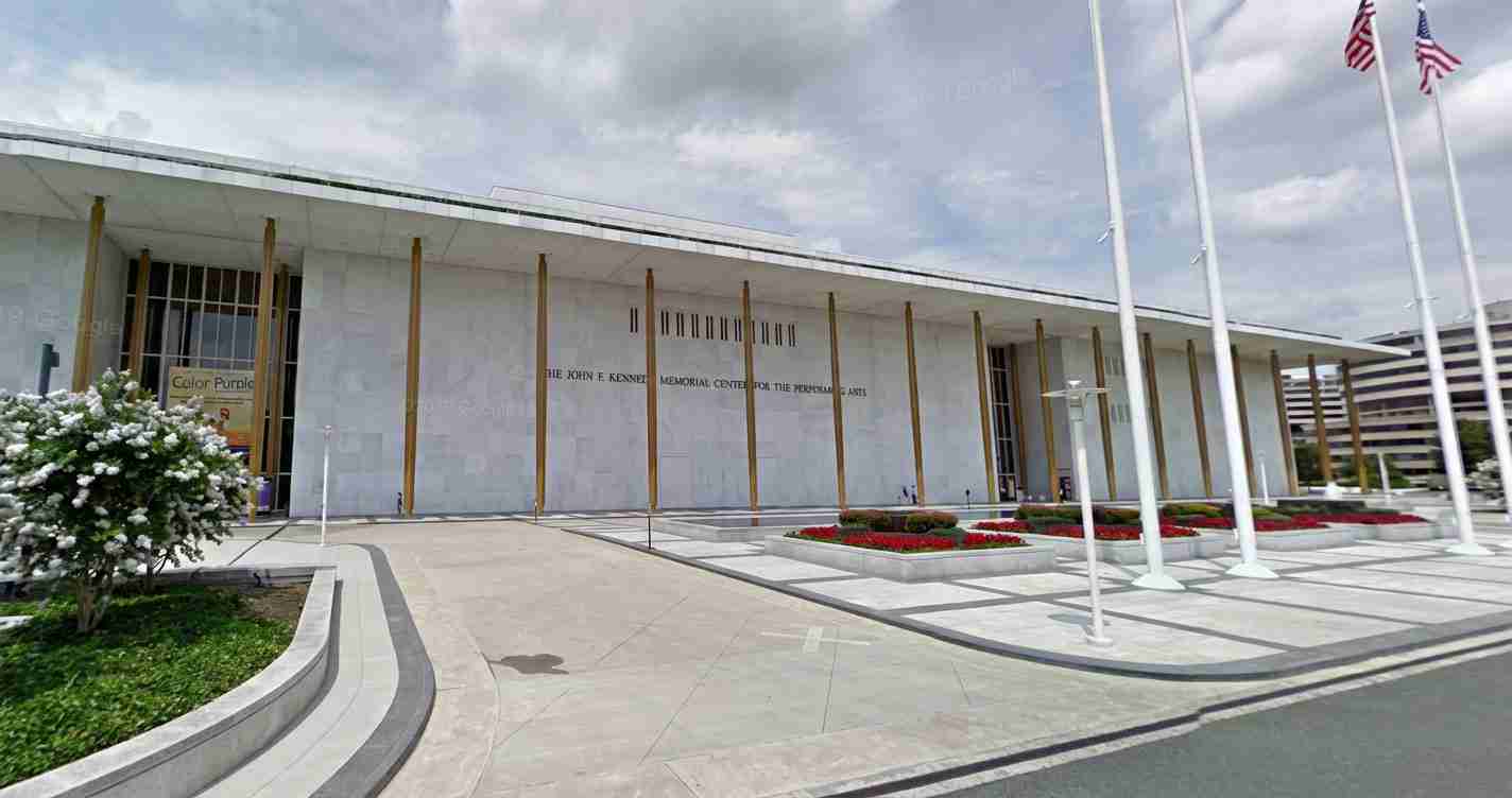 Modern memorial building with tall gold columns, flagpoles, and a landscaped front plaza under a cloudy sky.