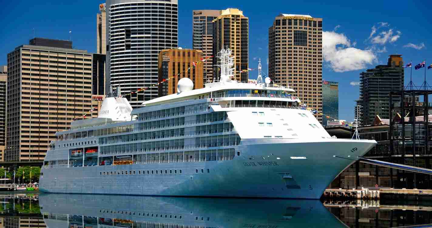 Large white cruise ship docked at a city harbor with tall modern buildings in the background, clear blue sky.