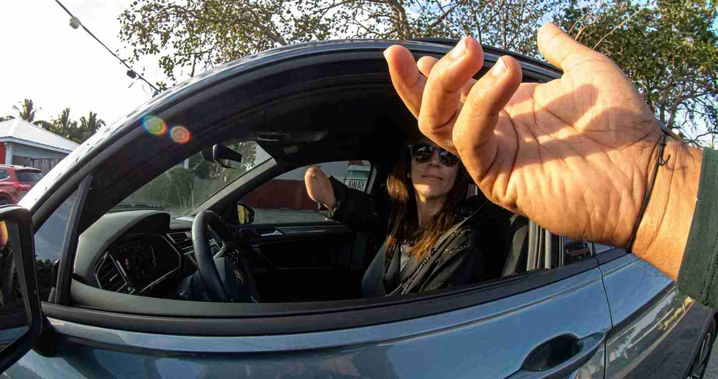Person sitting in car with window down, hand reaching toward the camera outside the vehicle in bright daylight. Lens flare visible.
