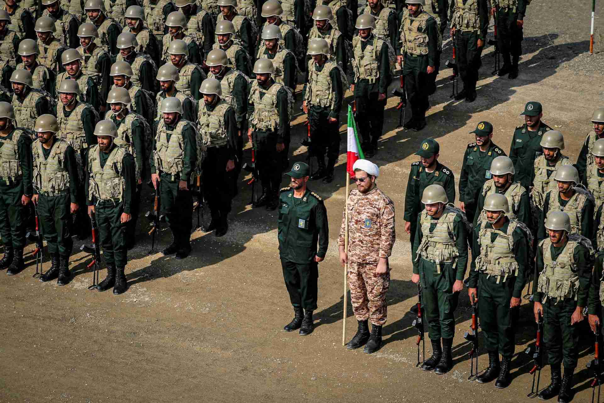 Large formation of soldiers in camouflage uniforms and helmets standing at attention on a dirt parade ground, with two officers and a flag bearer at the front.