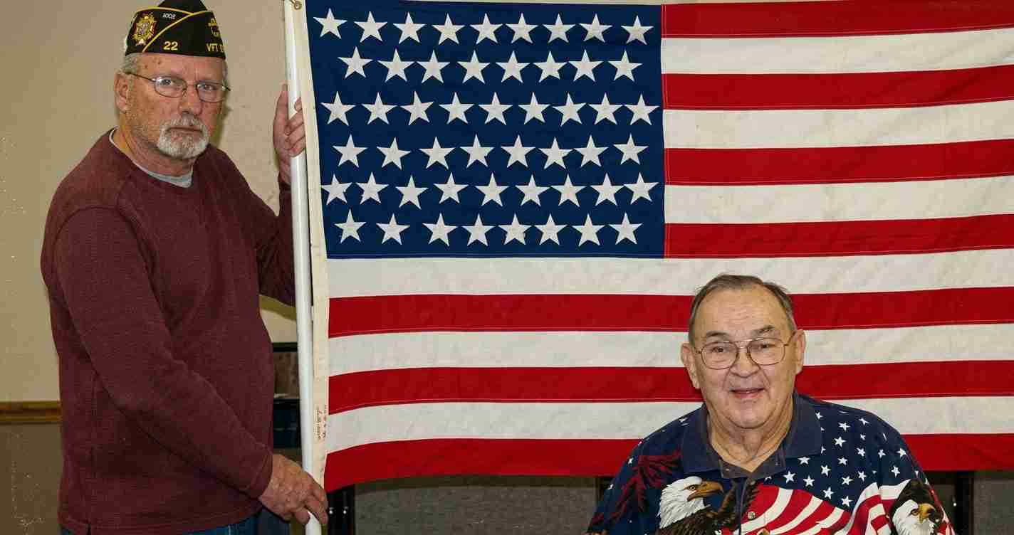 Two elderly men pose with a large American flag; one stands holding the flagpole, wearing a veteran cap and maroon sweater, the other sits in a patriotic shirt and smiles.