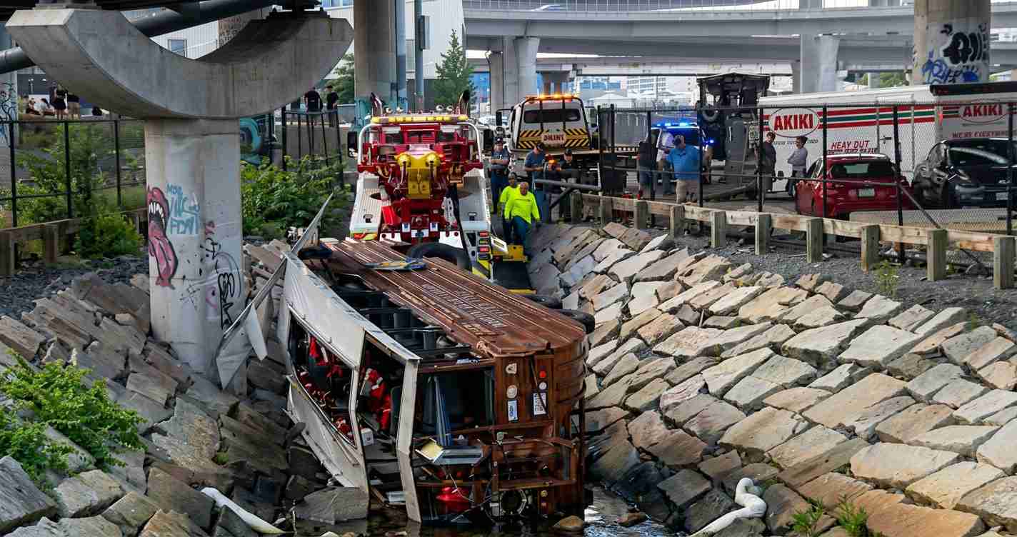Bus wrecked in a rock-lined drainage channel beneath elevated highways, surrounded by emergency responders and tow equipment.