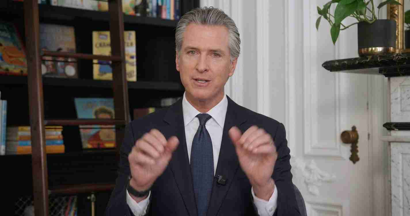 Man in a dark suit and tie speaks to the camera, gesturing with his hands in a room with bookshelves behind him.