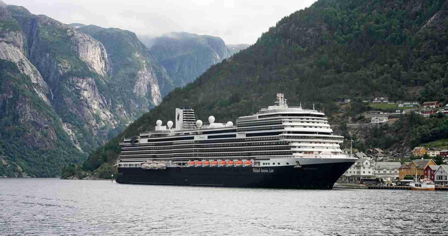 Large black-and-white cruise ship docked near a fjord with steep green mountains and a small village along the shore