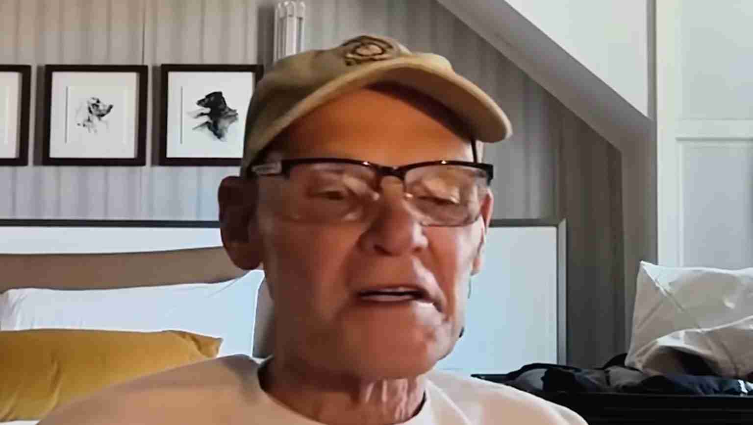 Older man wearing glasses and a tan cap speaks during a video call in a bedroom.