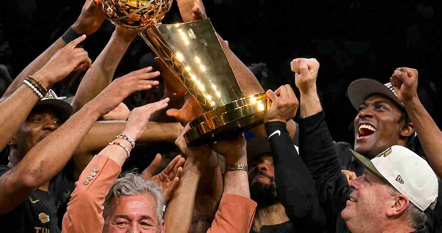 Team of basketball players celebrating victory, lifting a large gold trophy overhead with raised arms and joyful faces.