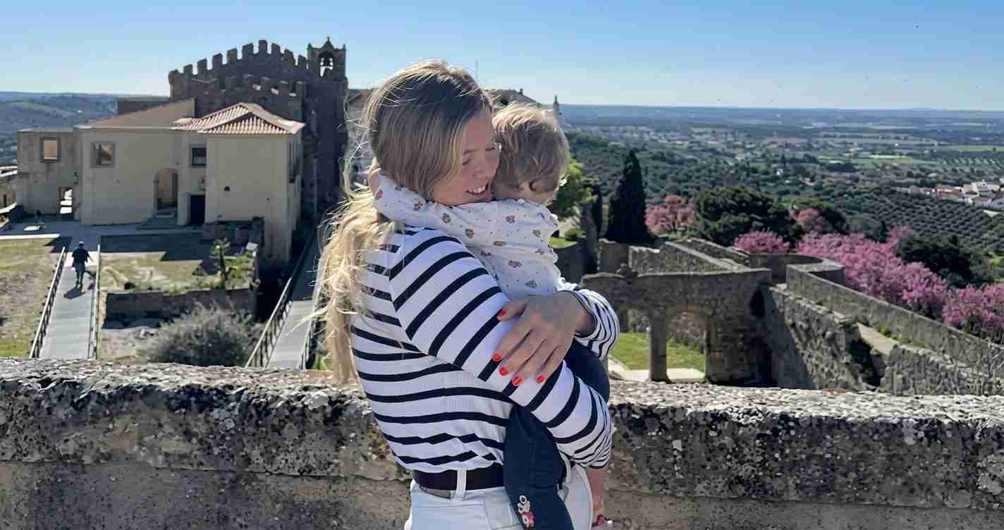 Woman in a striped shirt hugs a child on a stone wall with a medieval fortress and valley backdrop under a clear blue sky.