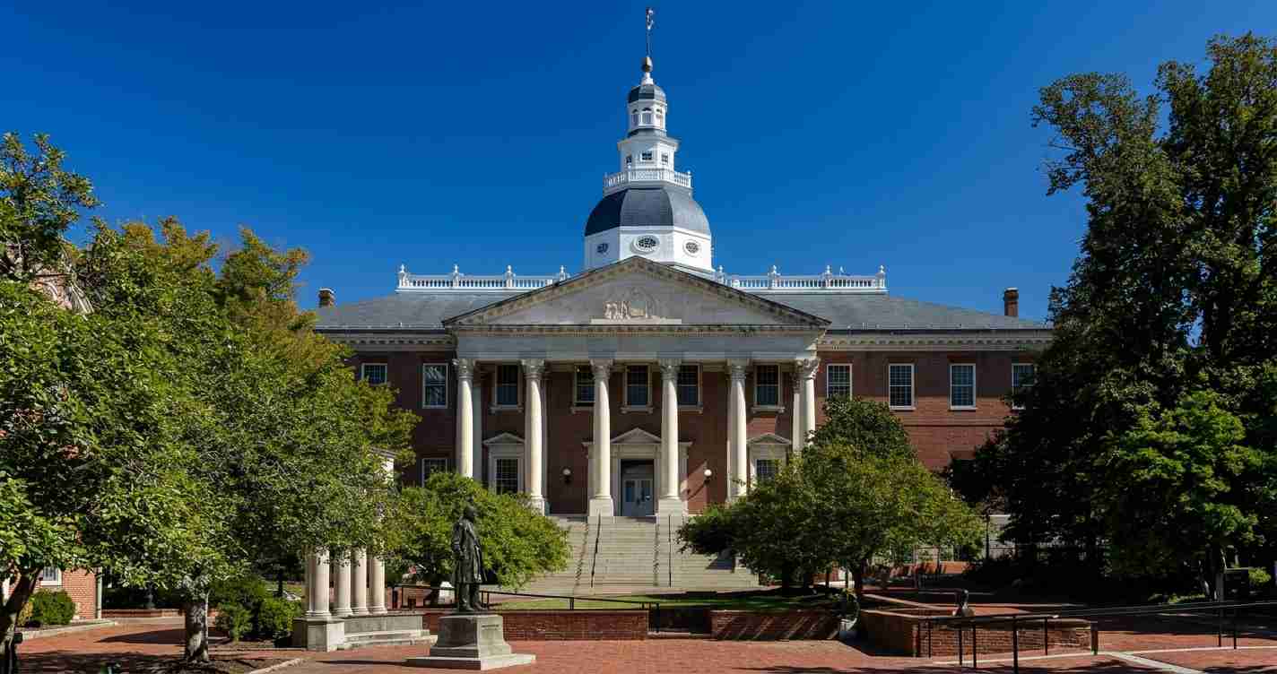 Front view of a neoclassical university building with tall white columns, a pediment, and a white-domed tower, set in a tree-filled plaza under a blue sky