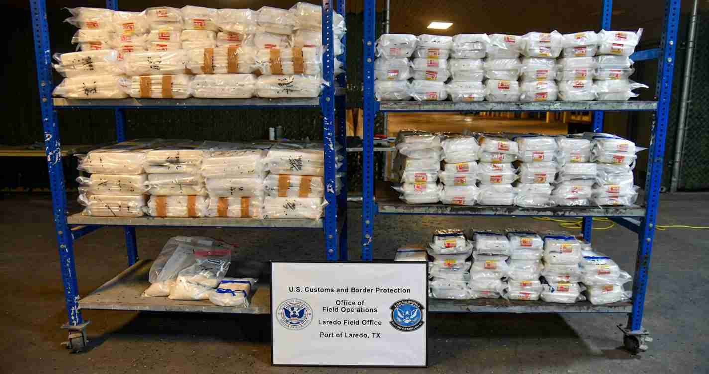 Stacks of white bags on blue metal shelving in a warehouse, with a CBP sign in front reading U.S. Customs and Border Protection, Port of Laredo, TX.