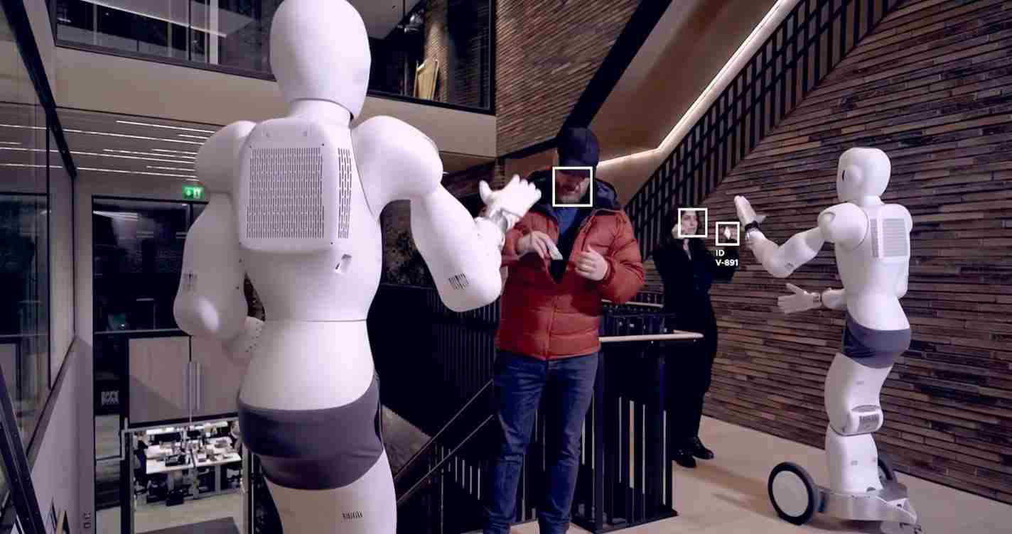 Two white humanoid robots high-five a man in a red jacket in a modern interior corridor