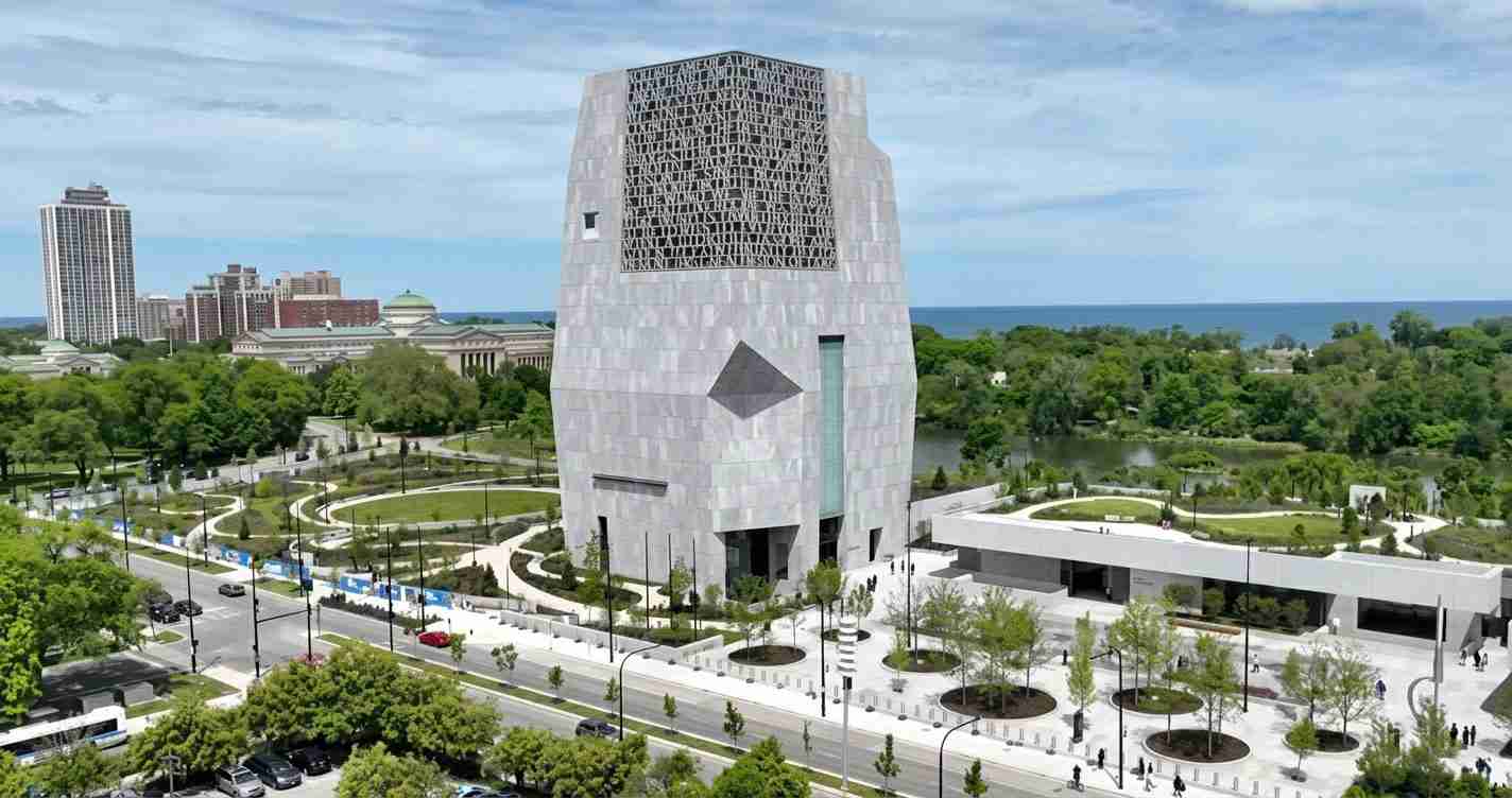 A tall modern gray tower with a lattice top sits in a landscaped park by a waterfront, seen from an aerial view.