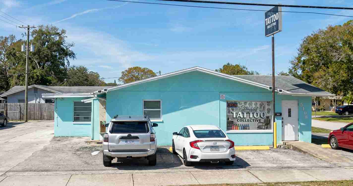 Turquoise single-story tattoo shop with a large window display reading 'Tattoo Collective' and two cars parked in front on a sunny street.