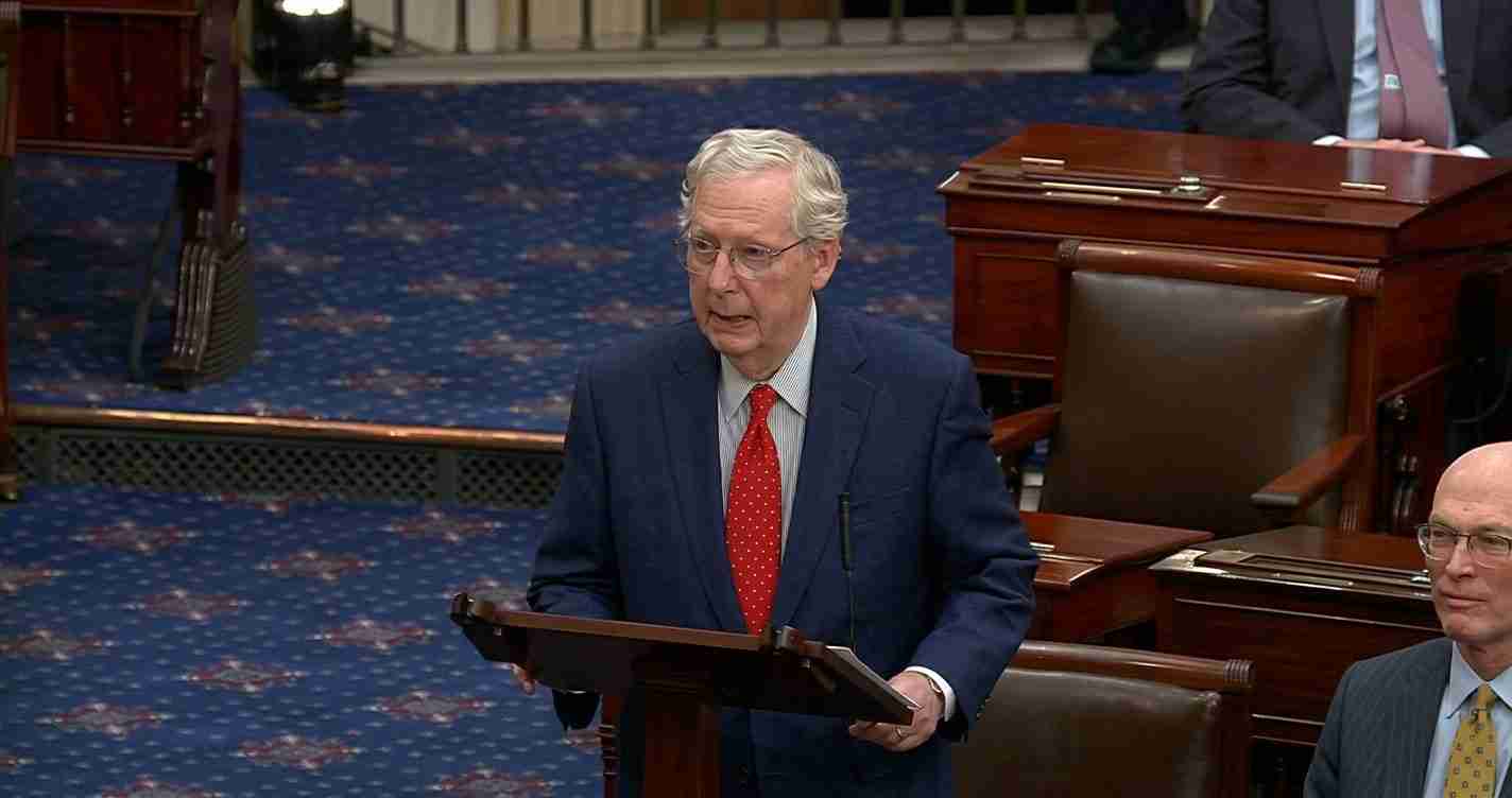 Older man in a navy suit and red tie speaks at a wooden podium in a legislative chamber.