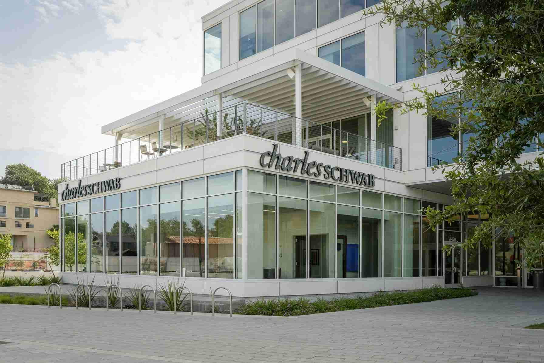Modern office building with glass walls and a visible Charles Schwab sign on the façade, sunny day with greenery nearby.