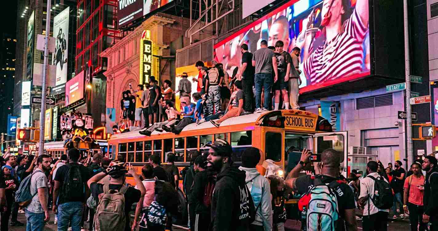 Crowded Times Square at night with a bright orange school bus topped by people and a sea of pedestrians around it, neon billboards in the background.