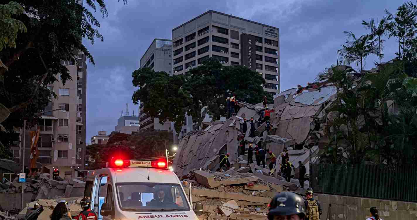 Emergency responders search through a collapsed building with rubble while an ambulance with flashing red lights is parked in the foreground.