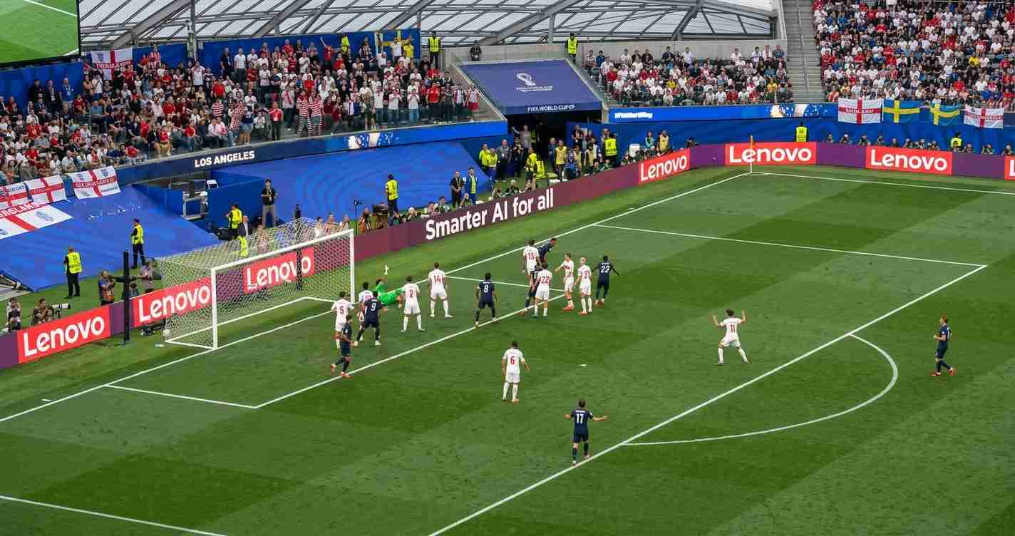 Soccer match action near the goal with players from two teams contesting a shot while a large crowd watches from the stands and advertising boards line the pitch edge.