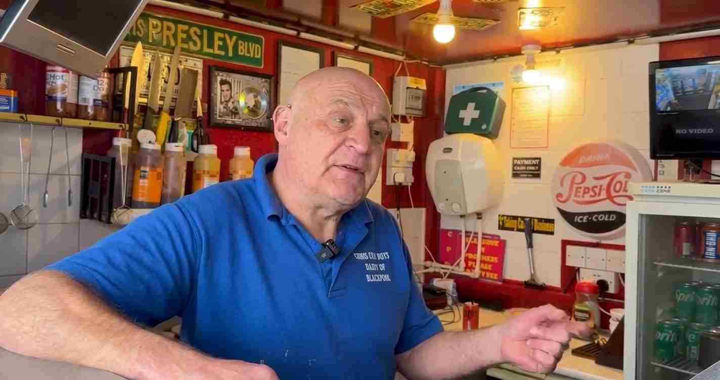 Man in a blue polo shirt behind a counter, talking, with shelves of bottles and retro signs in a colorful shop interior.