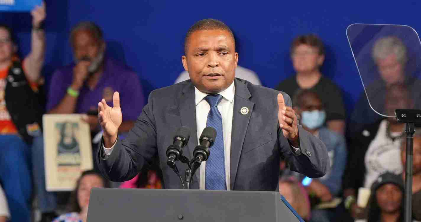 Man in a dark suit and blue tie speaks at a podium with two microphones, gesturing with his hands. 2) Politician delivering a speech at a public event, audience in the background. 3) Speaker at a rally addressing a crowd, microphones in front of him. 4) Formal press conference setup with microphones and podium as a man delivers an address. 5) Close-up of a man in a suit addressing an audience at a political event.