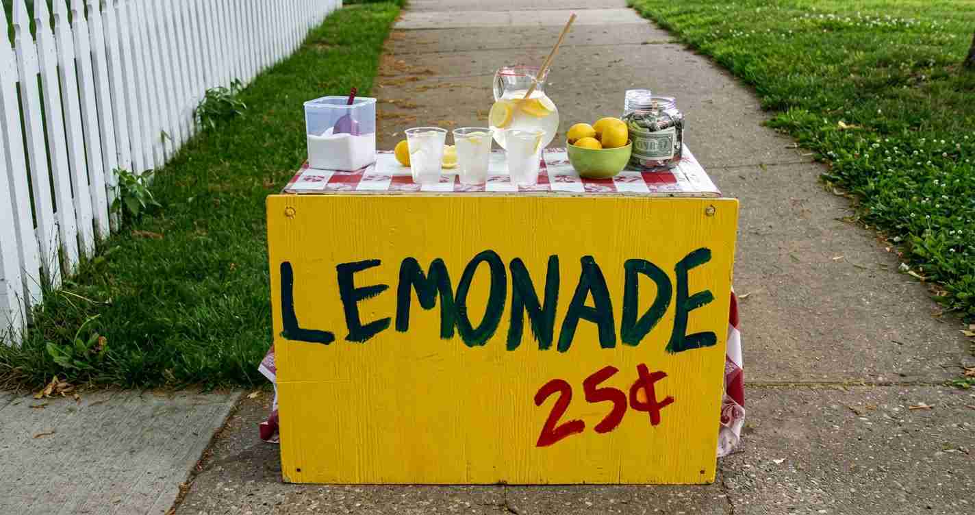 A yellow lemonade stand on a sidewalk with cups, a pitcher, lemons, and a jar of money, priced at 25 cents per cup.