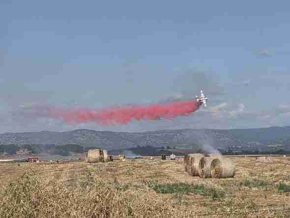 Crop-dusting airplane releasing red smoke over a field with round hay bales and distant hills under a blue sky