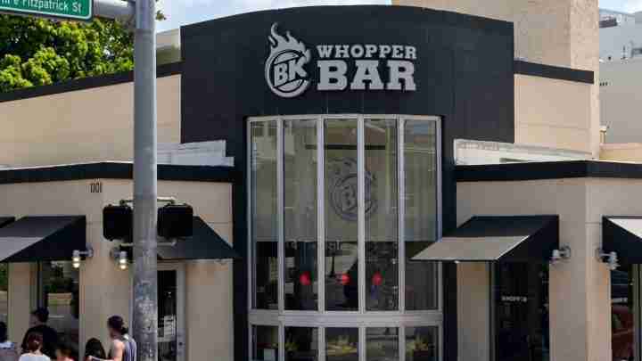 Whopper Bar storefront with a black façade, large curved glass entry, and the BK Whopper Bar sign above the door; people outside on a sunny day.
